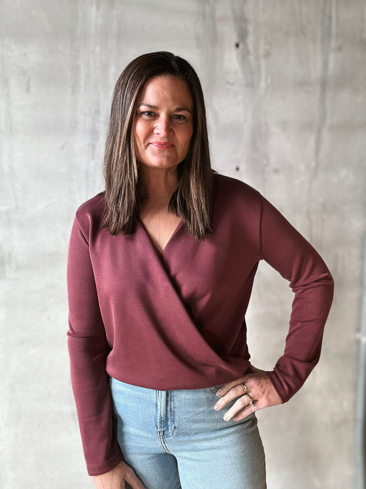 Woman wearing a maroon long-sleeve top and light blue jeans against a gray concrete wall.