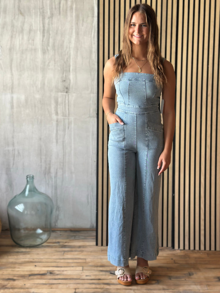 Woman wearing a light blue denim jumpsuit standing in front of a wooden wall with a large glass jar.