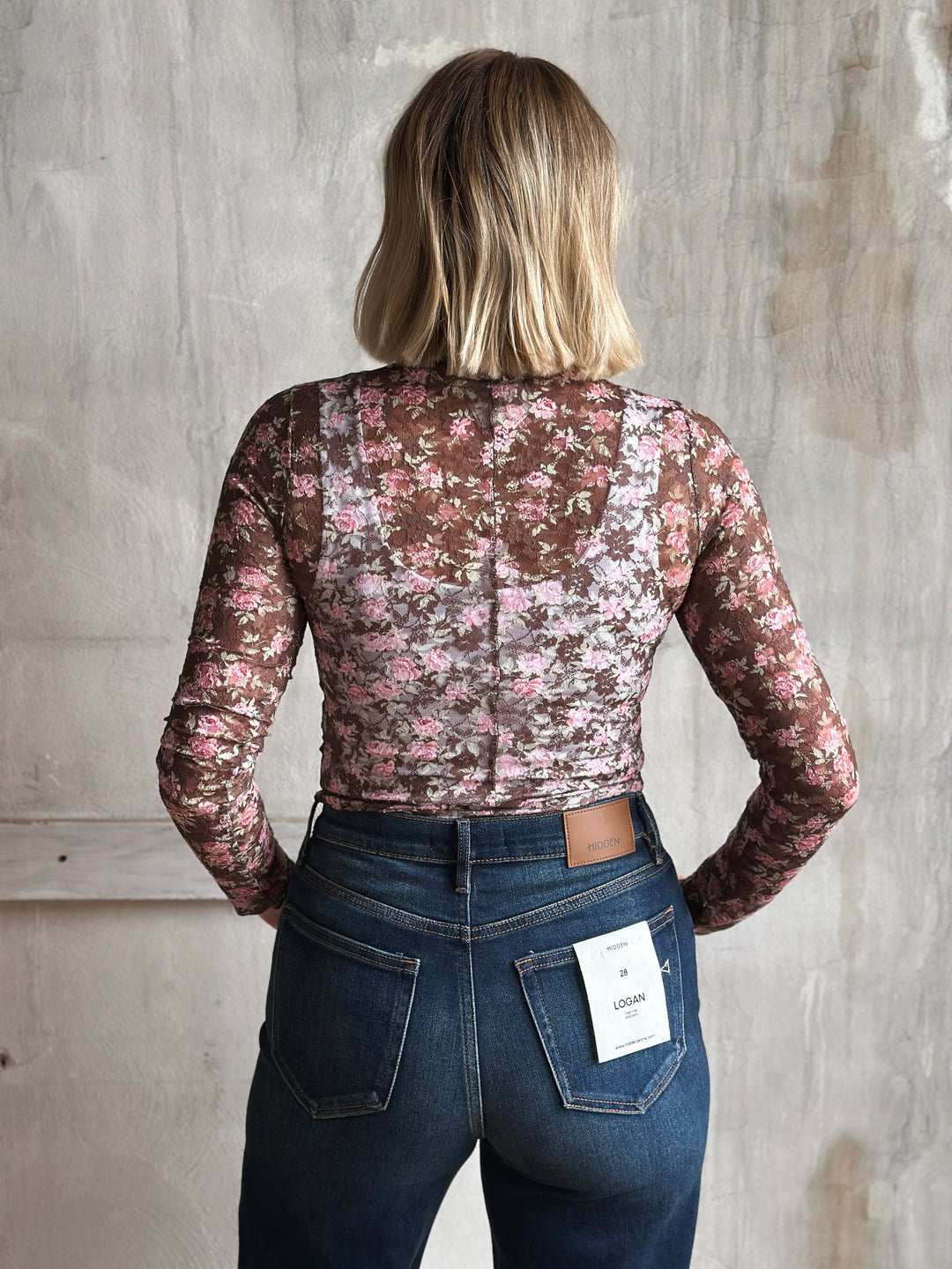 Person wearing a floral lace top and jeans with a visible tag against a plain background
