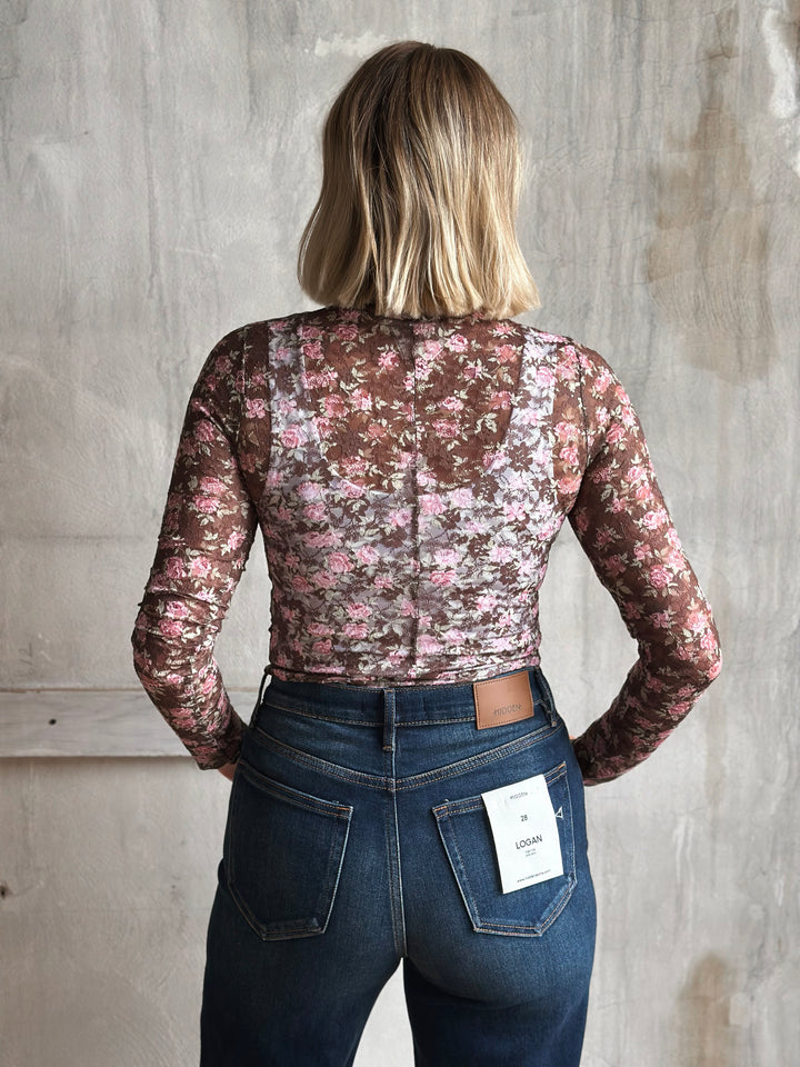 Person wearing a floral lace top and jeans with a visible tag against a plain background