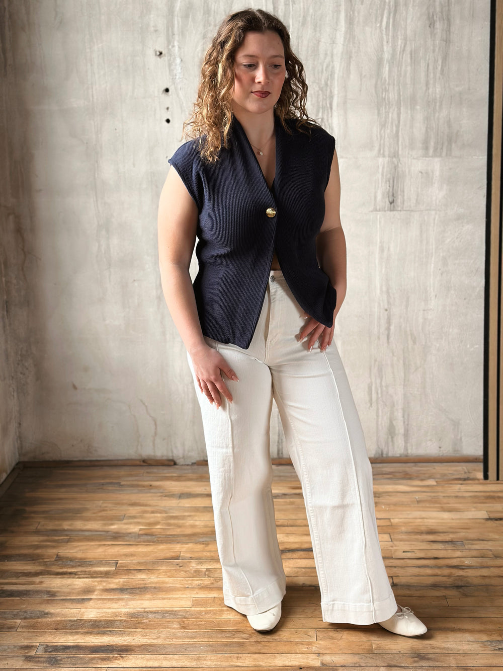 Woman wearing a navy sleeveless top and white jeans standing against a textured wall.