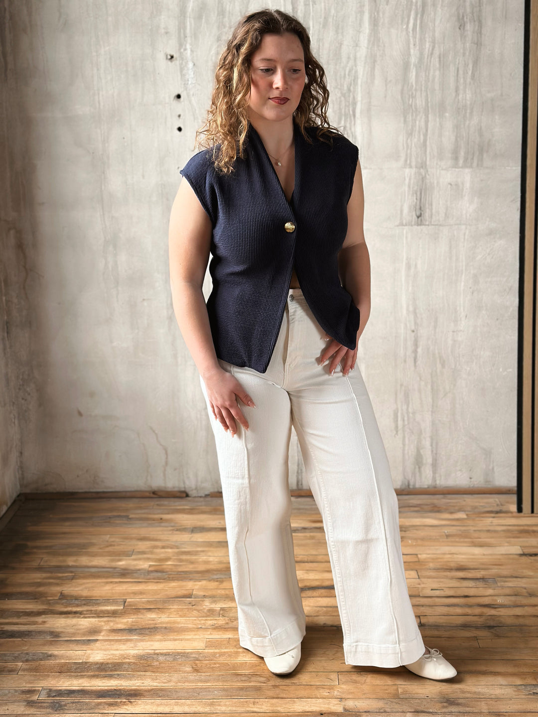 Woman wearing a navy sleeveless top and white jeans standing against a textured wall.