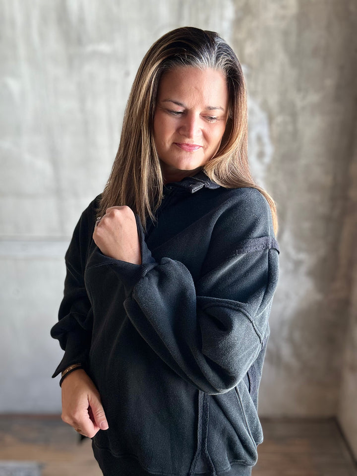 A woman with long brown hair wearing a dark charcoal gray oversized sweatshirt, standing against a textured concrete wall.