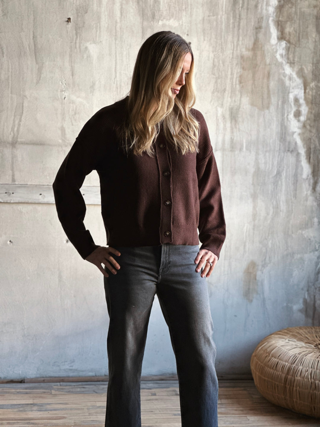 Woman wearing a brown cardigan and dark jeans standing against a textured wall.