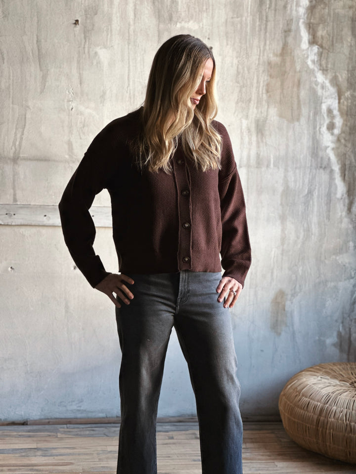 Woman wearing a brown cardigan and dark jeans standing against a textured wall.