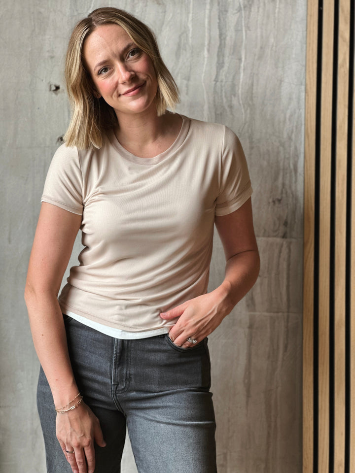Woman wearing a beige t-shirt and gray jeans standing against a wooden wall.