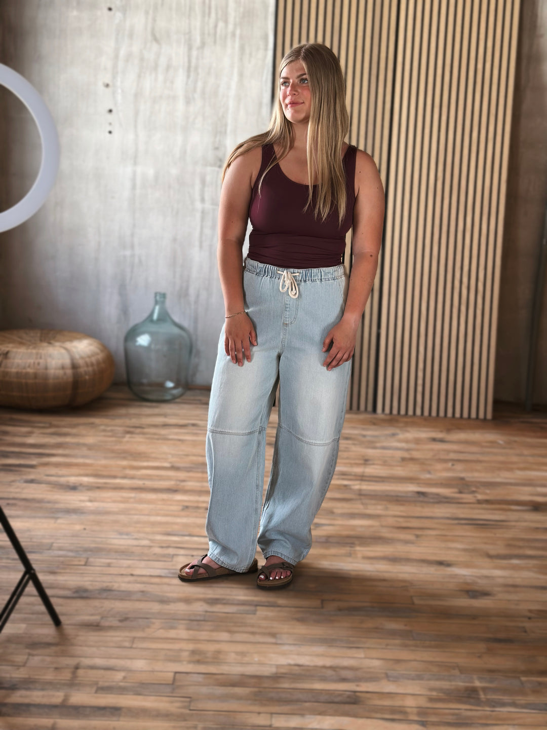Woman wearing a brown tank top and light blue jeans standing in a room with wooden flooring and a textured wall.