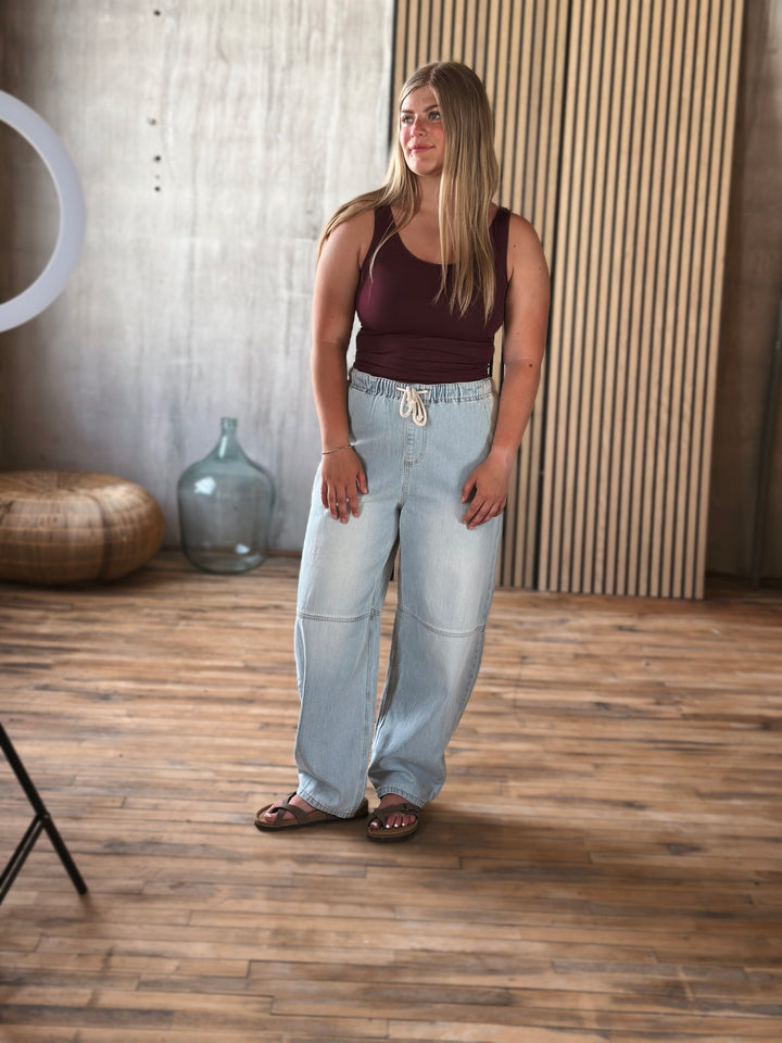 Woman wearing a brown tank top and light blue jeans standing in a room with wooden flooring and a textured wall.