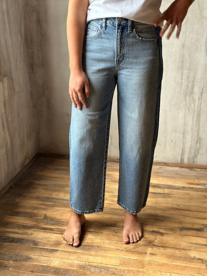 A person wearing light-wash blue cropped jeans with a white top, standing barefoot on a wooden floor against a textured concrete wall.