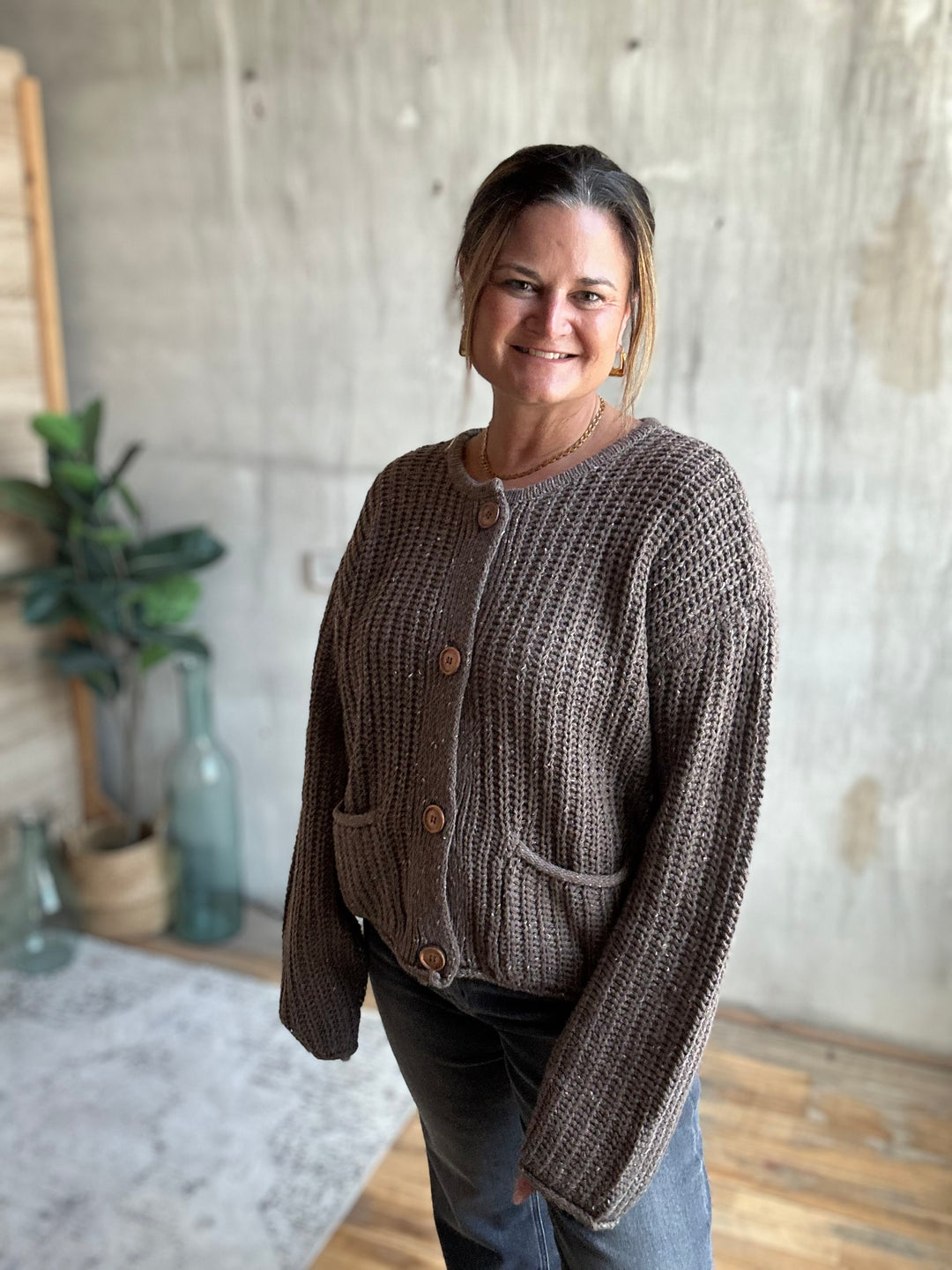 A woman wearing a brown chunky knit button-up cardigan with front pockets, standing in a room with a concrete wall background.