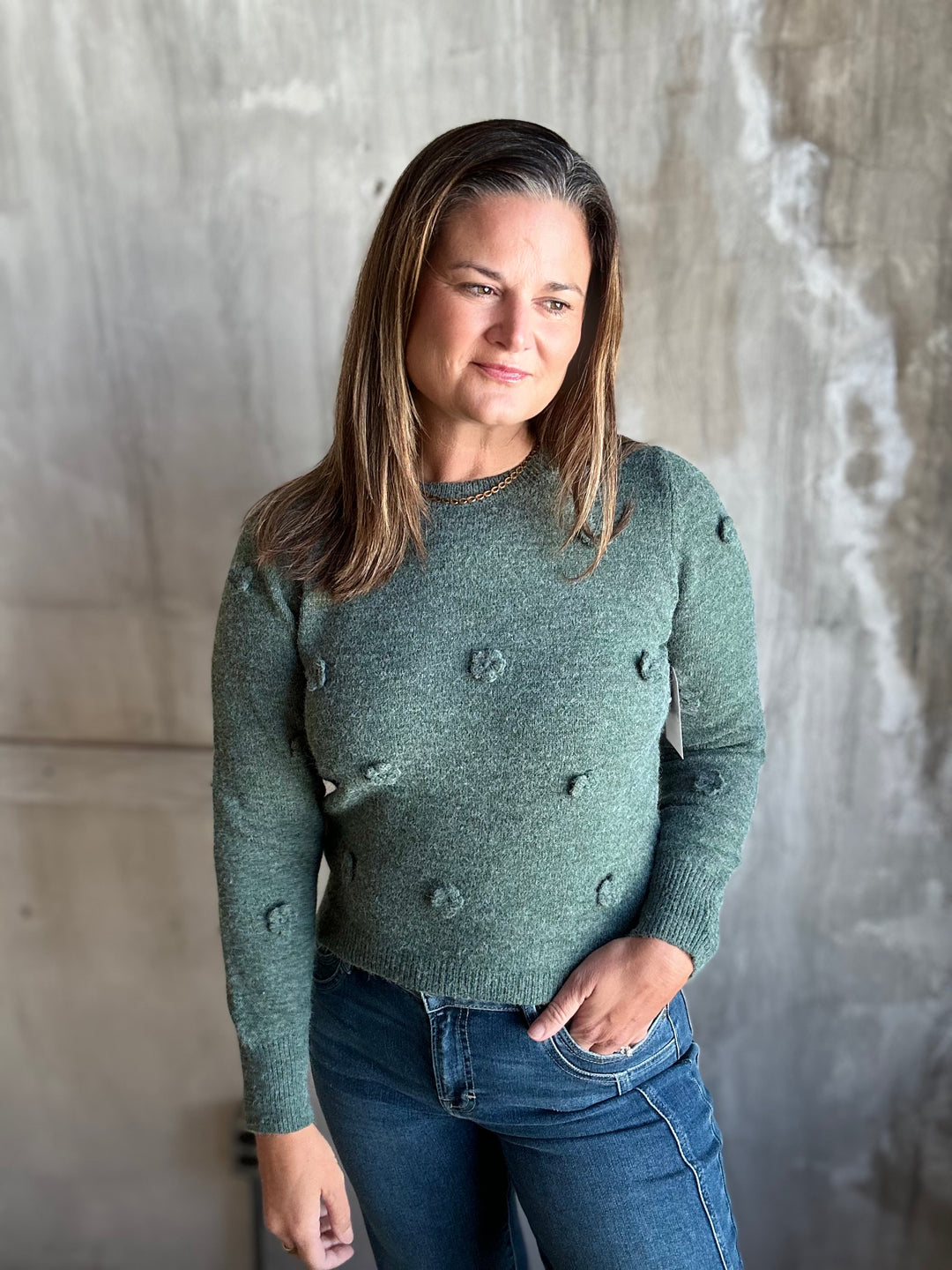 A woman wearing a sage green knit sweater with textured pom-pom details and blue jeans, standing against a textured concrete wall.