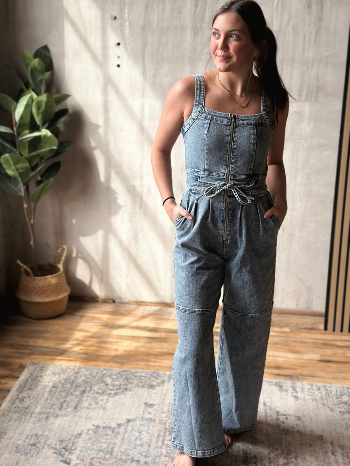 Woman wearing a denim jumpsuit standing in a room with a plant and rug.