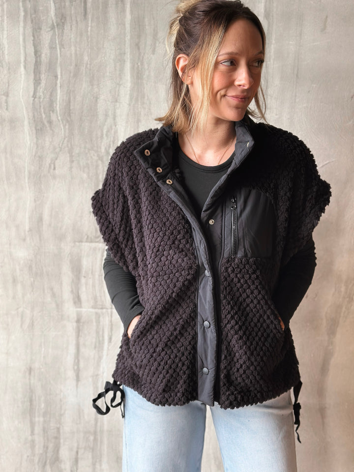 Woman wearing a black quilted vest over a gray shirt against a light wooden panel background
