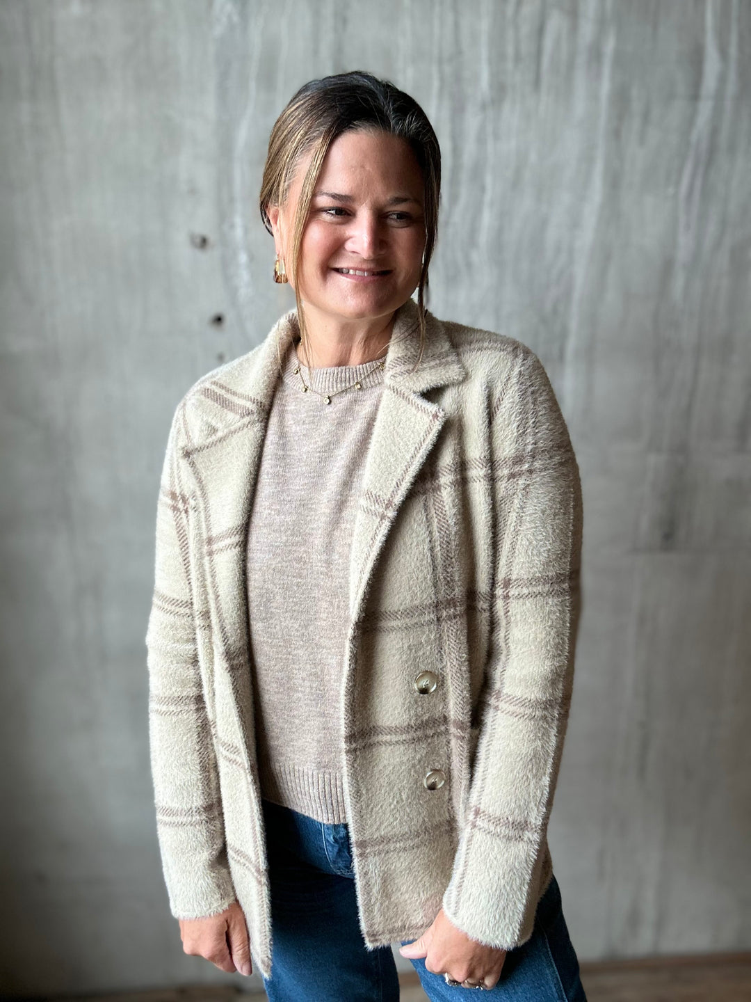 A woman wearing a cream and taupe plaid button-front cardigan over a beige knit sweater and blue jeans, standing against a textured gray concrete wall.