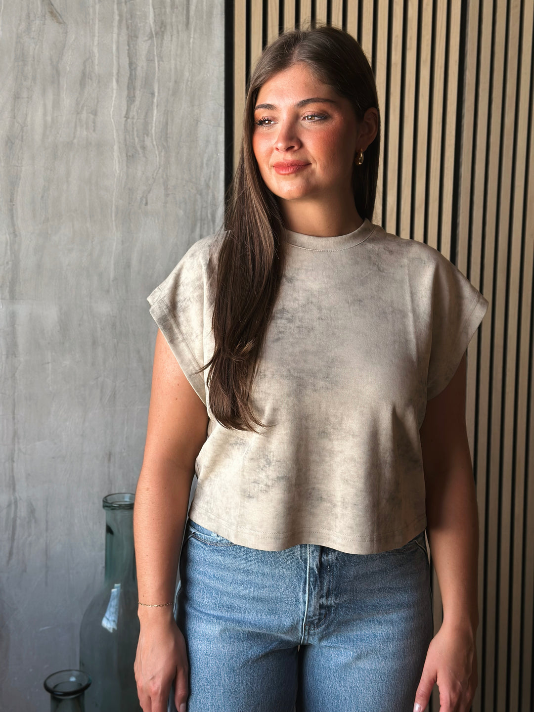 Woman wearing a beige top and blue jeans standing against a textured wall.