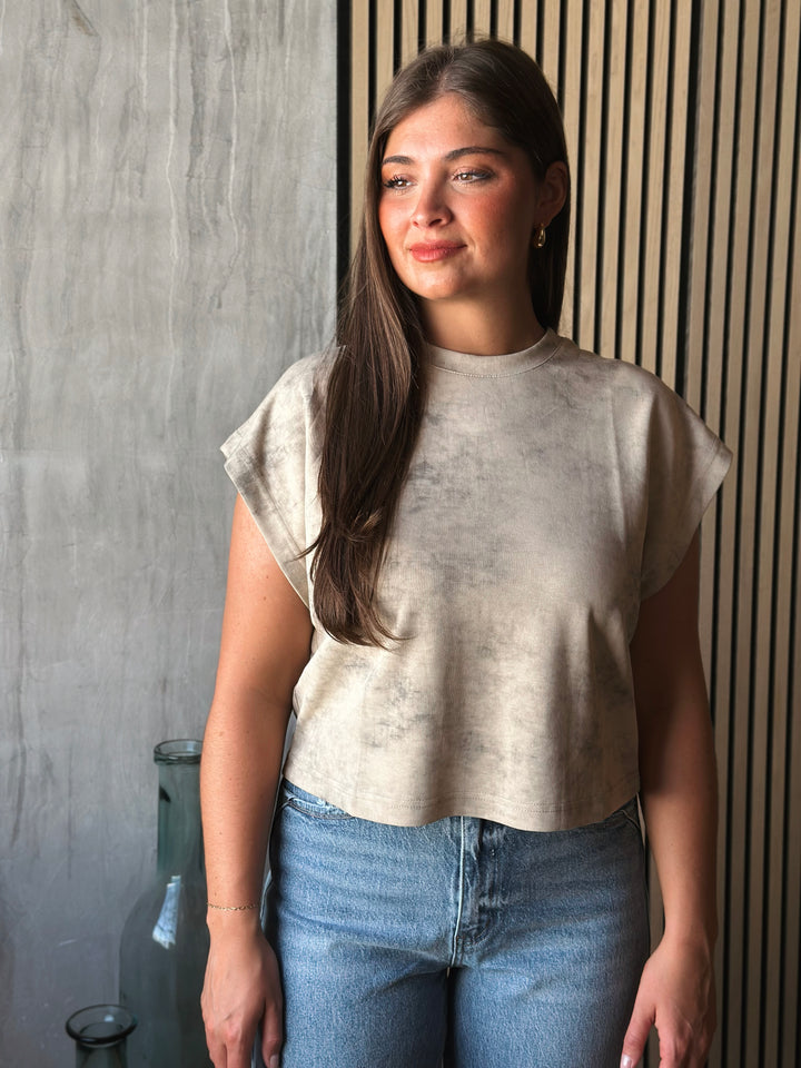 Woman wearing a beige top and blue jeans standing against a textured wall.