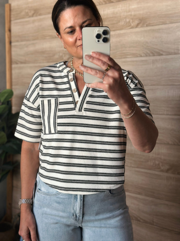 A woman wearing a short sleeved black and white striped v-neck shirt with buttons. With a pocket on the left side of the shirt. 