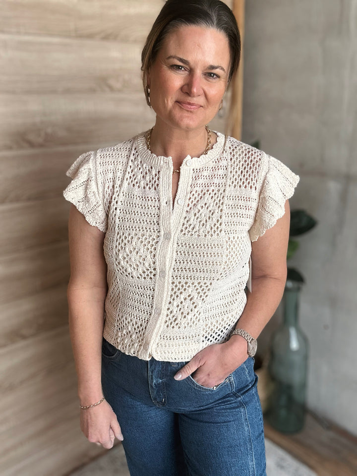 Woman wearing a white crochet top and blue jeans standing indoors.