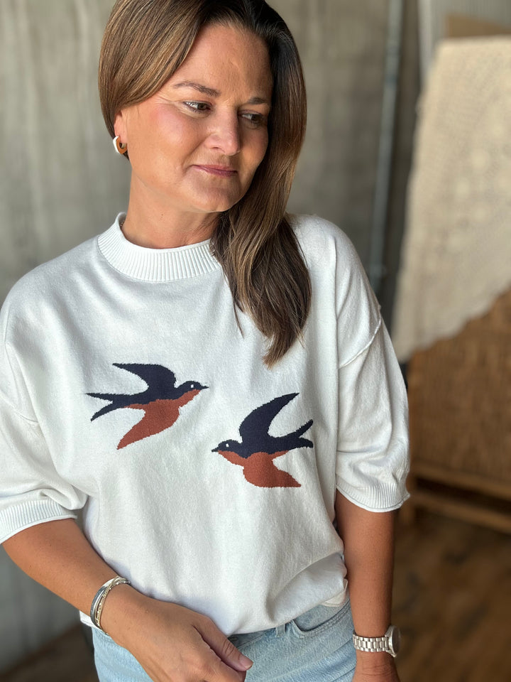 A woman wearing a white short-sleeved sweater with two embroidered swallow birds on the front, paired with light blue jeans.