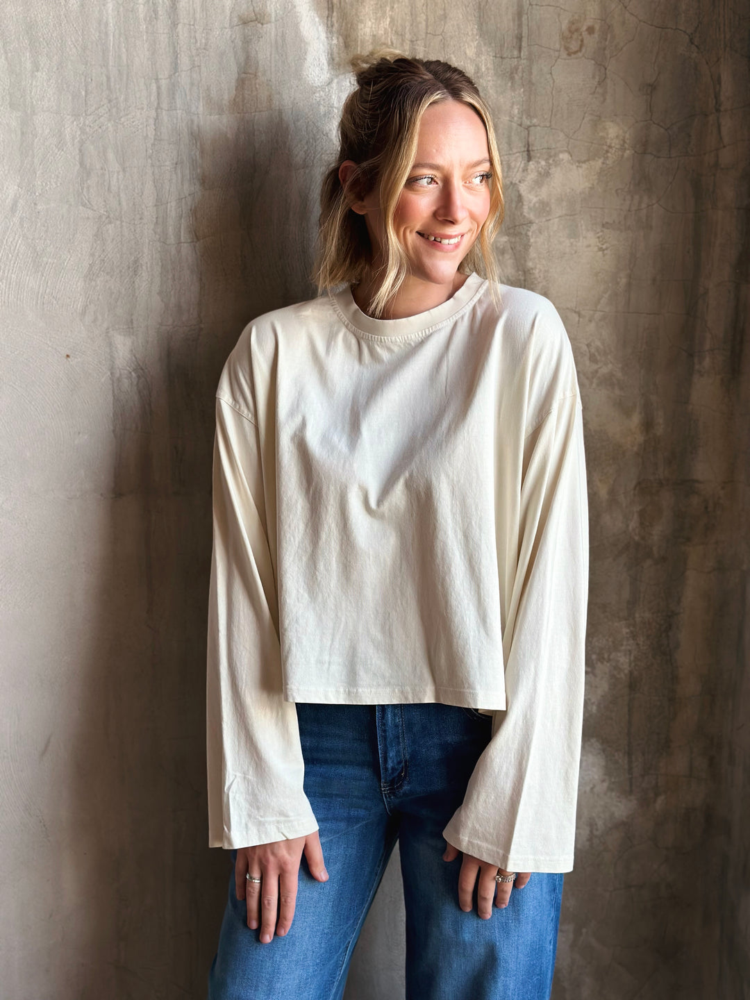 Woman wearing a white long-sleeve top and blue jeans against a textured wall.