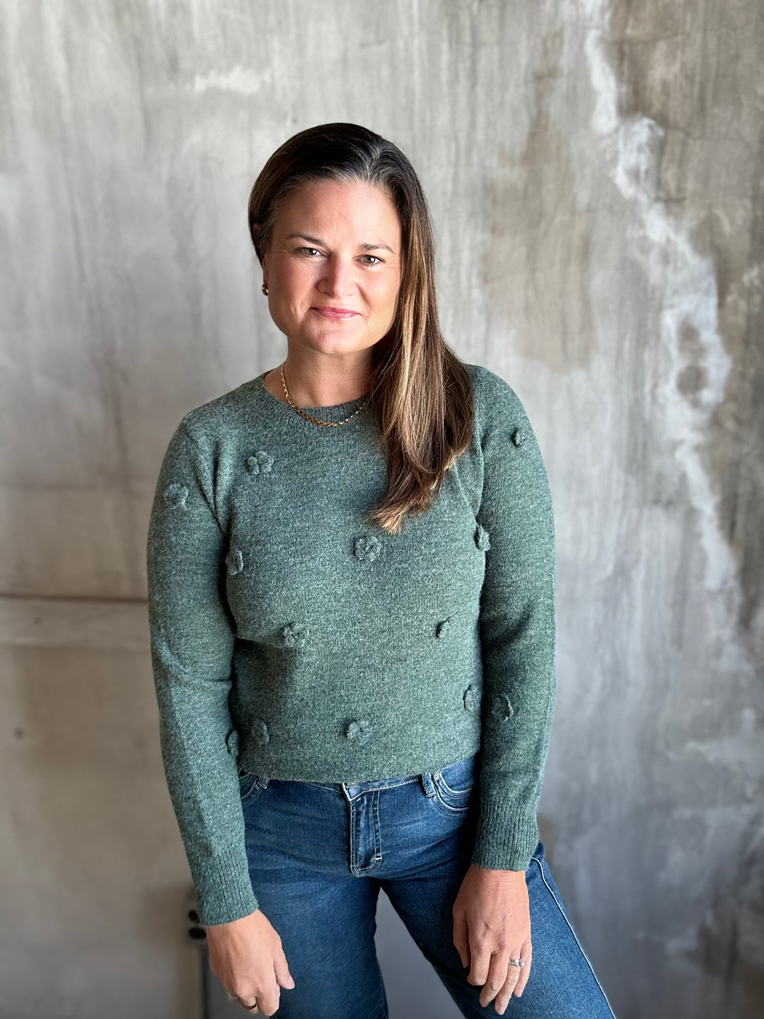 A woman wearing a sage green knit sweater with raised textured details and blue jeans, standing against a textured concrete wall.