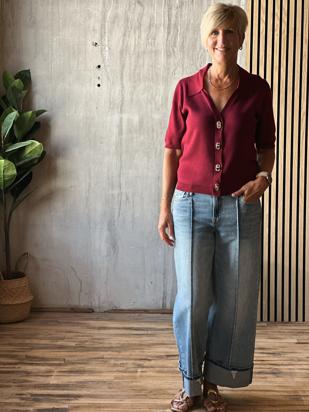 Woman wearing a red cardigan and blue jeans standing indoors with a plant and wooden floor in the background