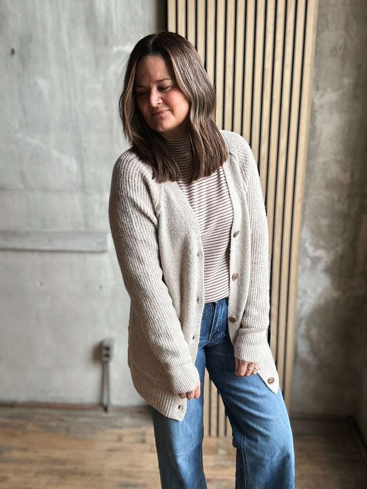 Woman wearing a beige cardigan over a striped shirt with blue jeans, standing against a textured wall.