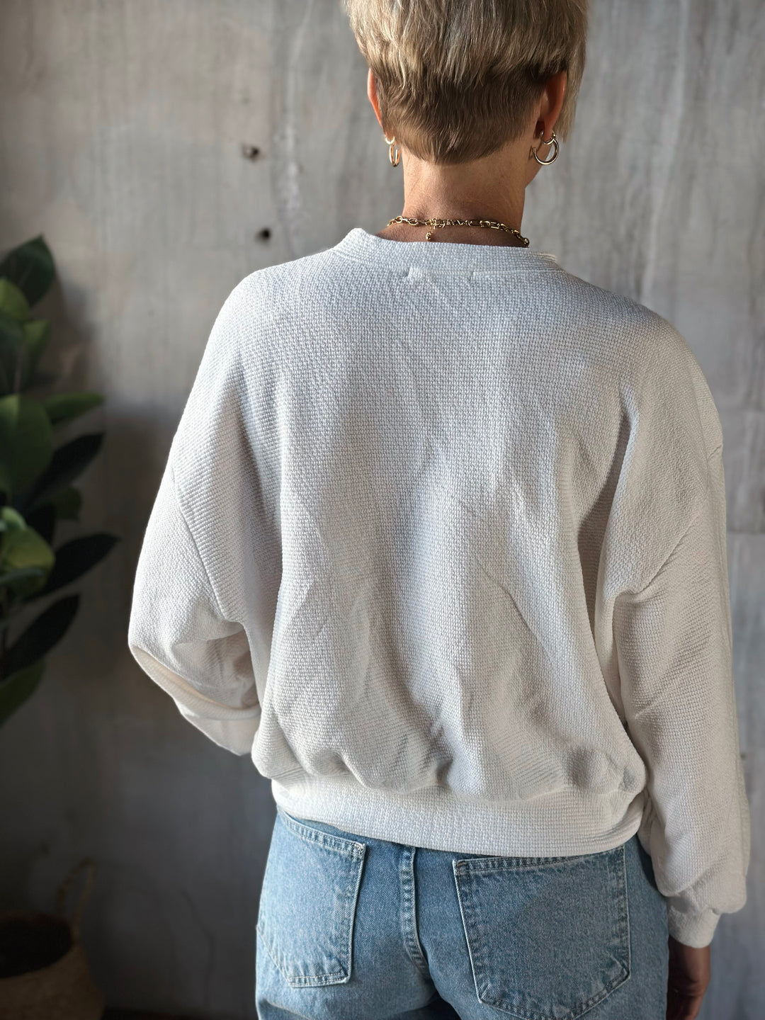 Person wearing a light gray sweater and blue jeans standing against a neutral wall.