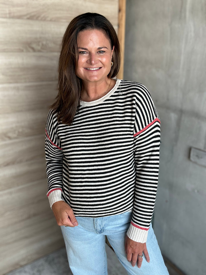 A woman wearing a black and white horizontally striped long-sleeve sweater with thin pink accent stripes on the shoulders and cuffs, paired with light blue jeans, standing against a split wood and concrete wall.