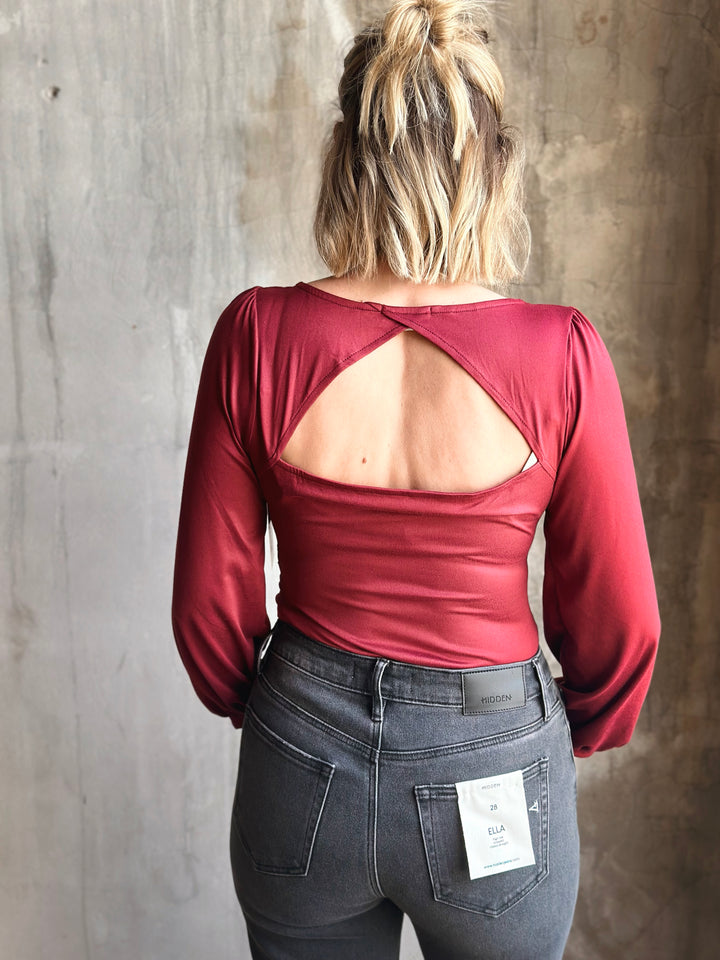 Person wearing a red long-sleeve top with an open back and dark jeans against a textured wall.