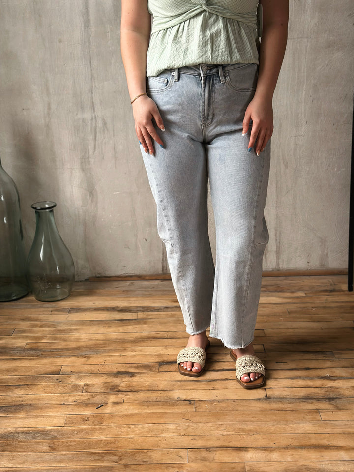 Person wearing light blue jeans and a green top standing in a room with wooden flooring and a concrete wall.
