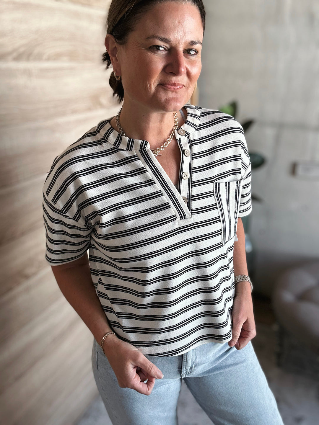 A woman wearing a short sleeved black and white striped v-neck shirt with buttons. With a pocket on the left side of the shirt. 