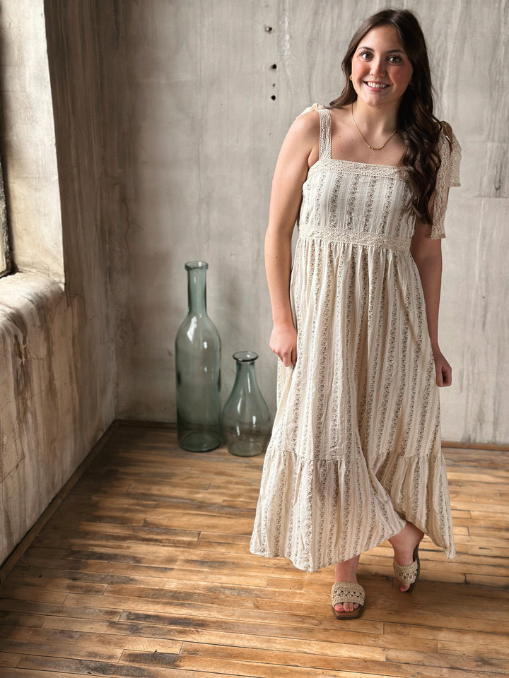 Woman in a white dress with floral designs standing in a room with wooden floor and concrete wall.