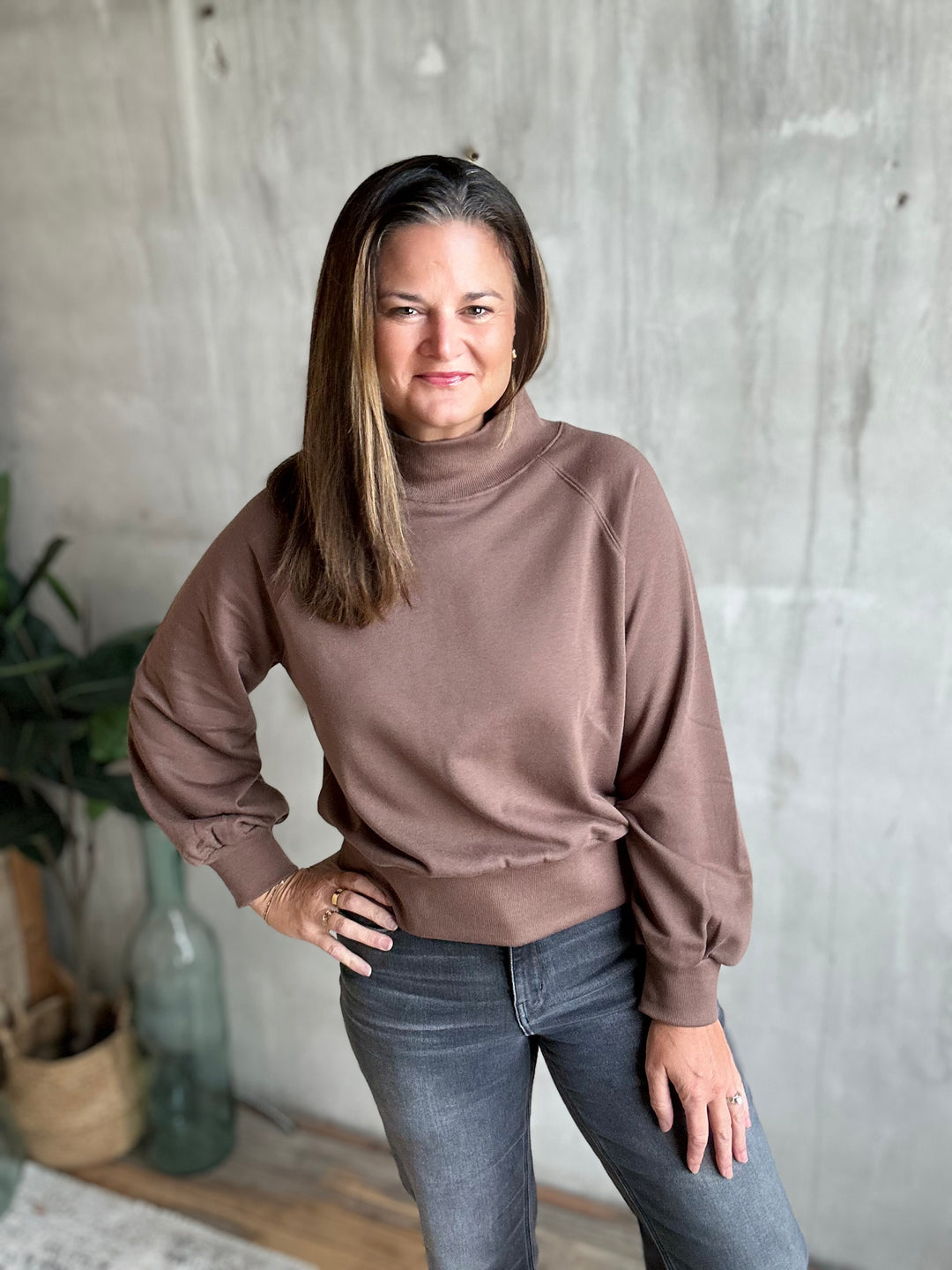 A woman wearing a brown mock neck sweatshirt and gray jeans, standing against a textured concrete wall.