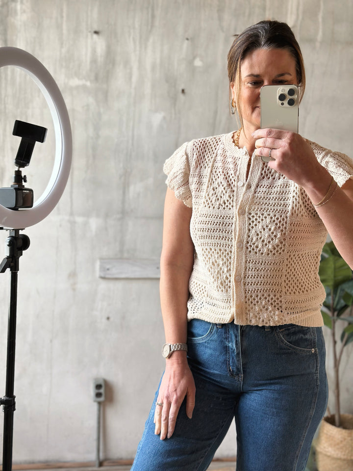 Woman taking a mirror selfie wearing a beige crochet top and blue jeans.