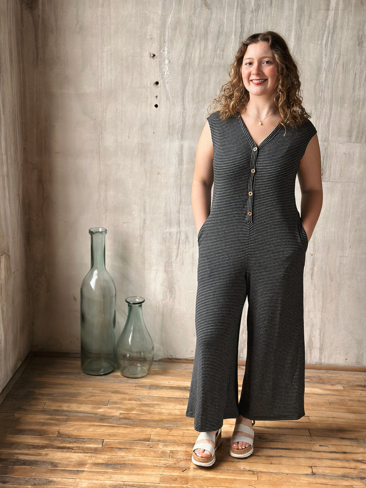 Woman wearing a black striped jumpsuit standing in a room with wooden floor and concrete wall.
