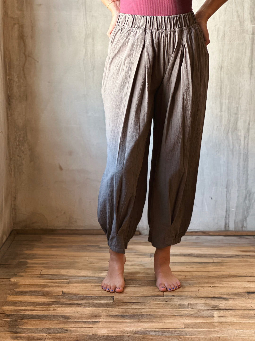 Person wearing brown wide-leg pants standing in a room with wooden floor and textured walls.