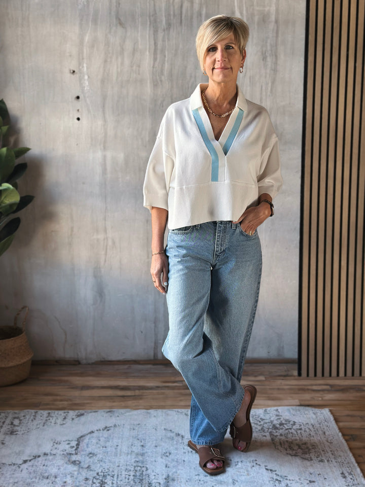 Woman wearing a white blouse with blue accents and blue jeans standing in a room with wooden flooring and a plant.