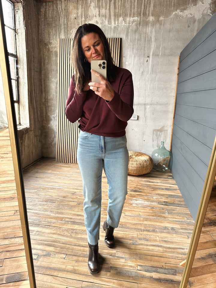 Woman taking a mirror selfie wearing a burgundy sweater and light blue jeans in a room with wooden floor and concrete wall.