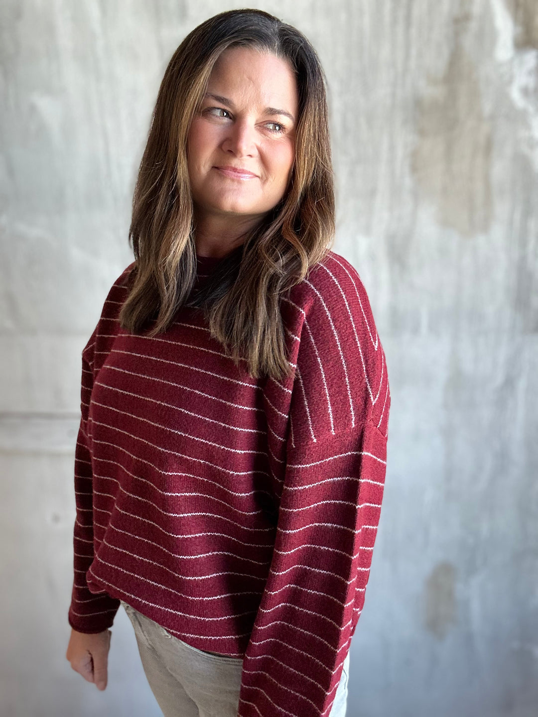 A woman wearing a maroon knit sweater with thin white horizontal stripes, paired with light beige pants, standing against a textured gray concrete wall.