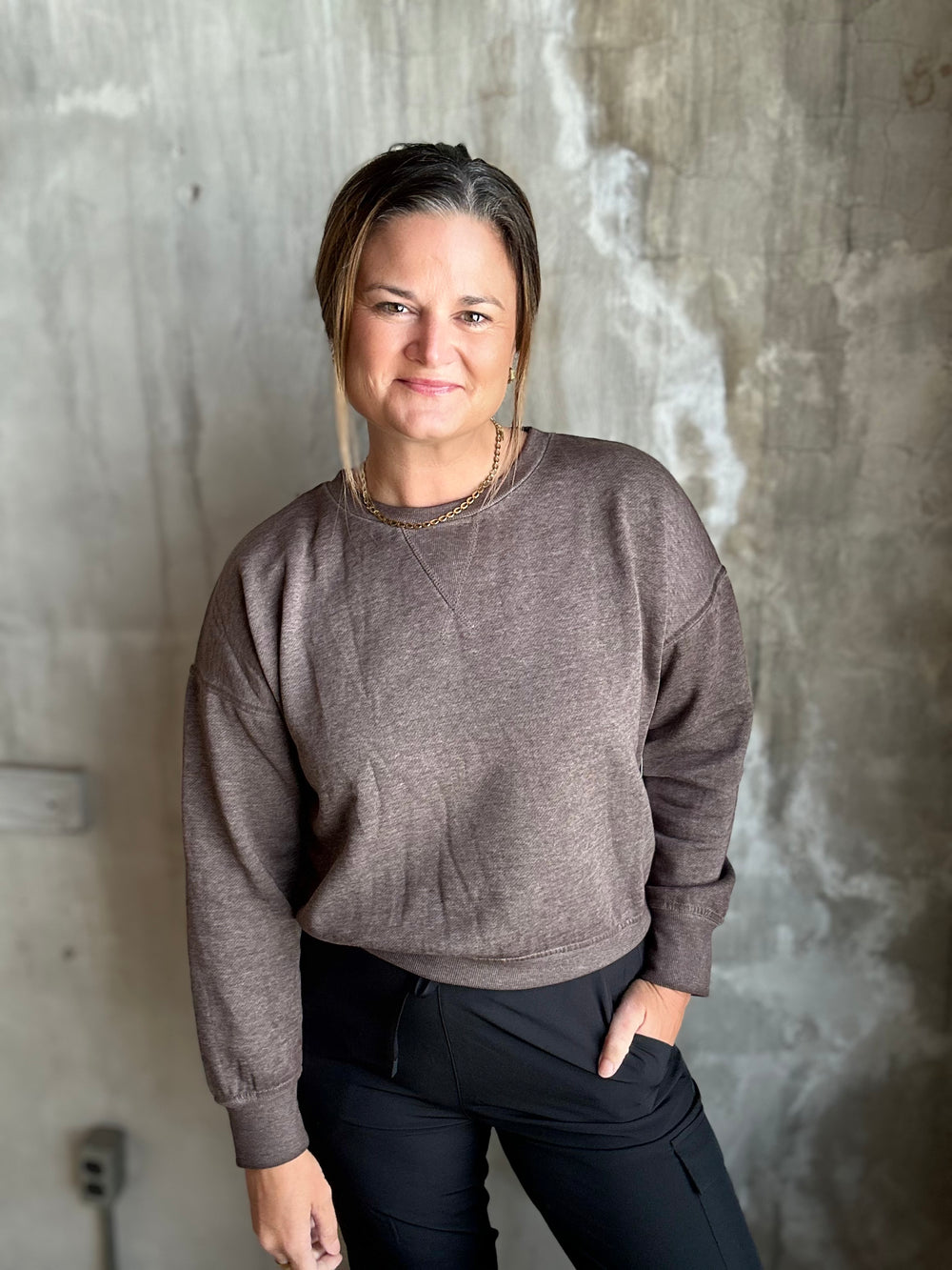 A woman wearing a heathered taupe-brown crewneck sweatshirt and black pants, standing against a textured concrete wall.
