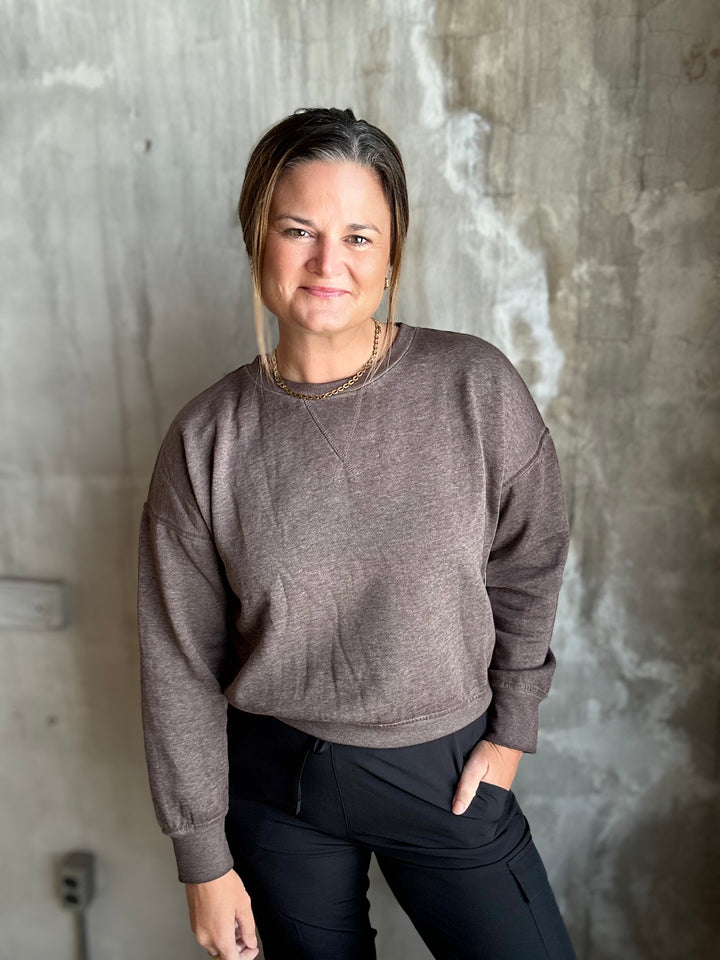 A woman wearing a heathered taupe-brown crewneck sweatshirt and black pants, standing against a textured concrete wall.
