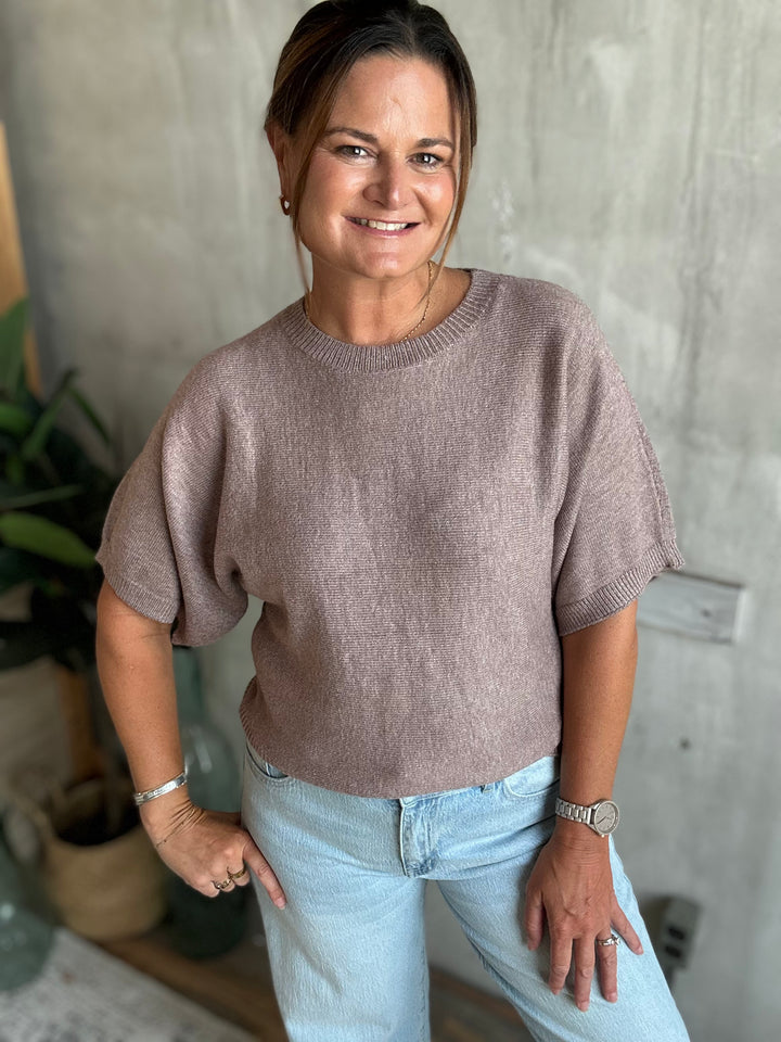 A woman wearing a short-sleeve knit sweater in a dusty mauve-brown color, paired with light-wash blue jeans.