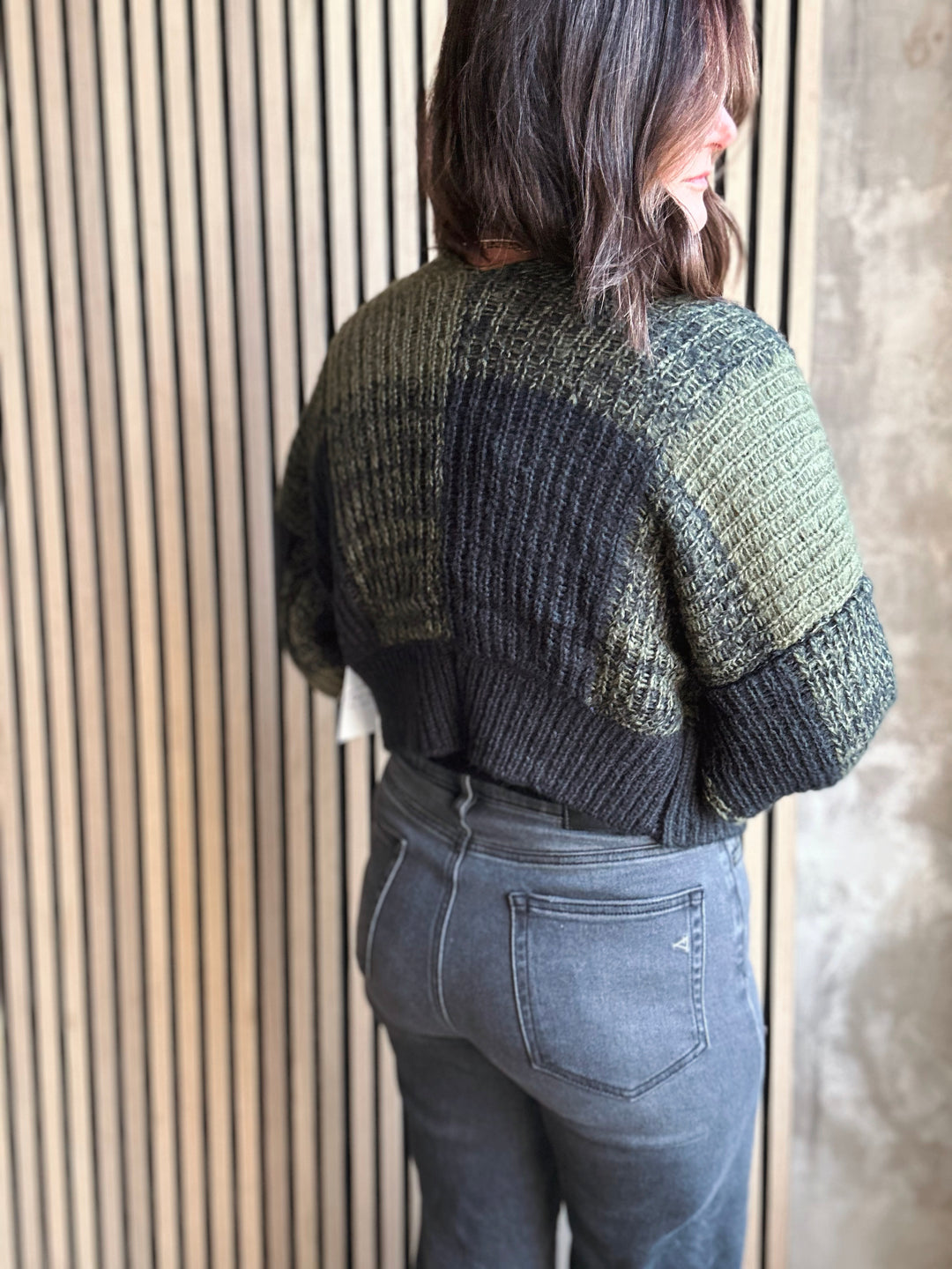 Person wearing a green and black knitted sweater and gray jeans against a wooden paneled wall.