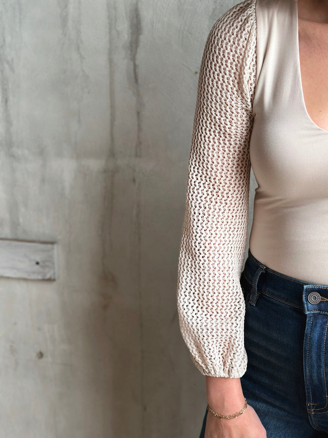 Person wearing a beige knit long-sleeve top against a concrete wall.