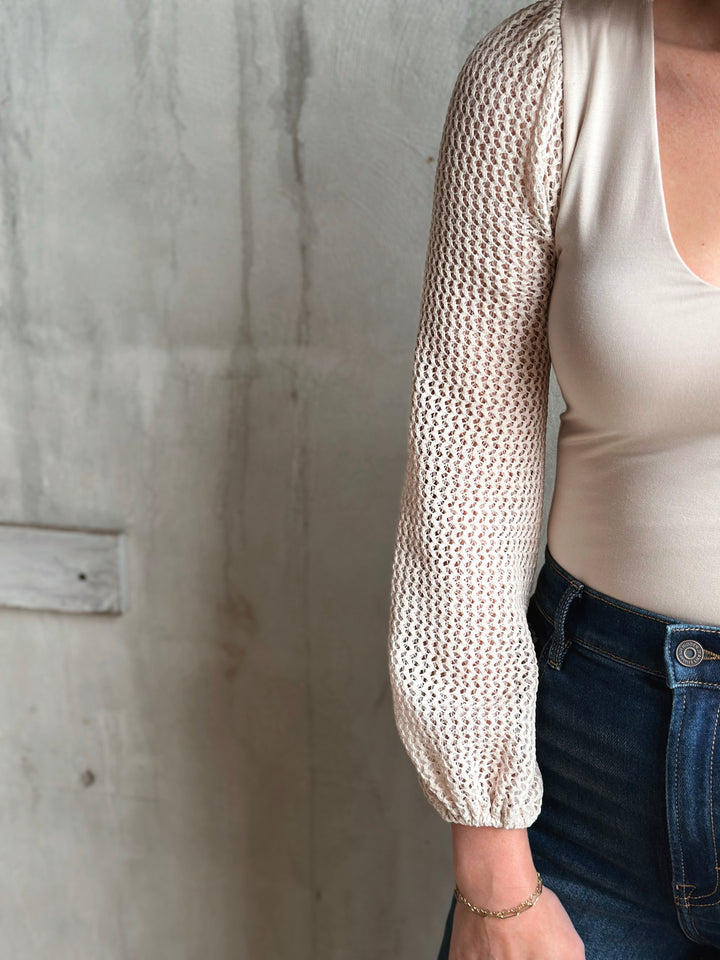Person wearing a beige knit long-sleeve top against a concrete wall.