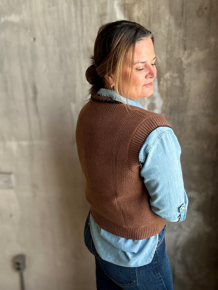 A woman wearing a brown ribbed knit vest over a light blue denim shirt and dark blue jeans, standing against a textured concrete wall.