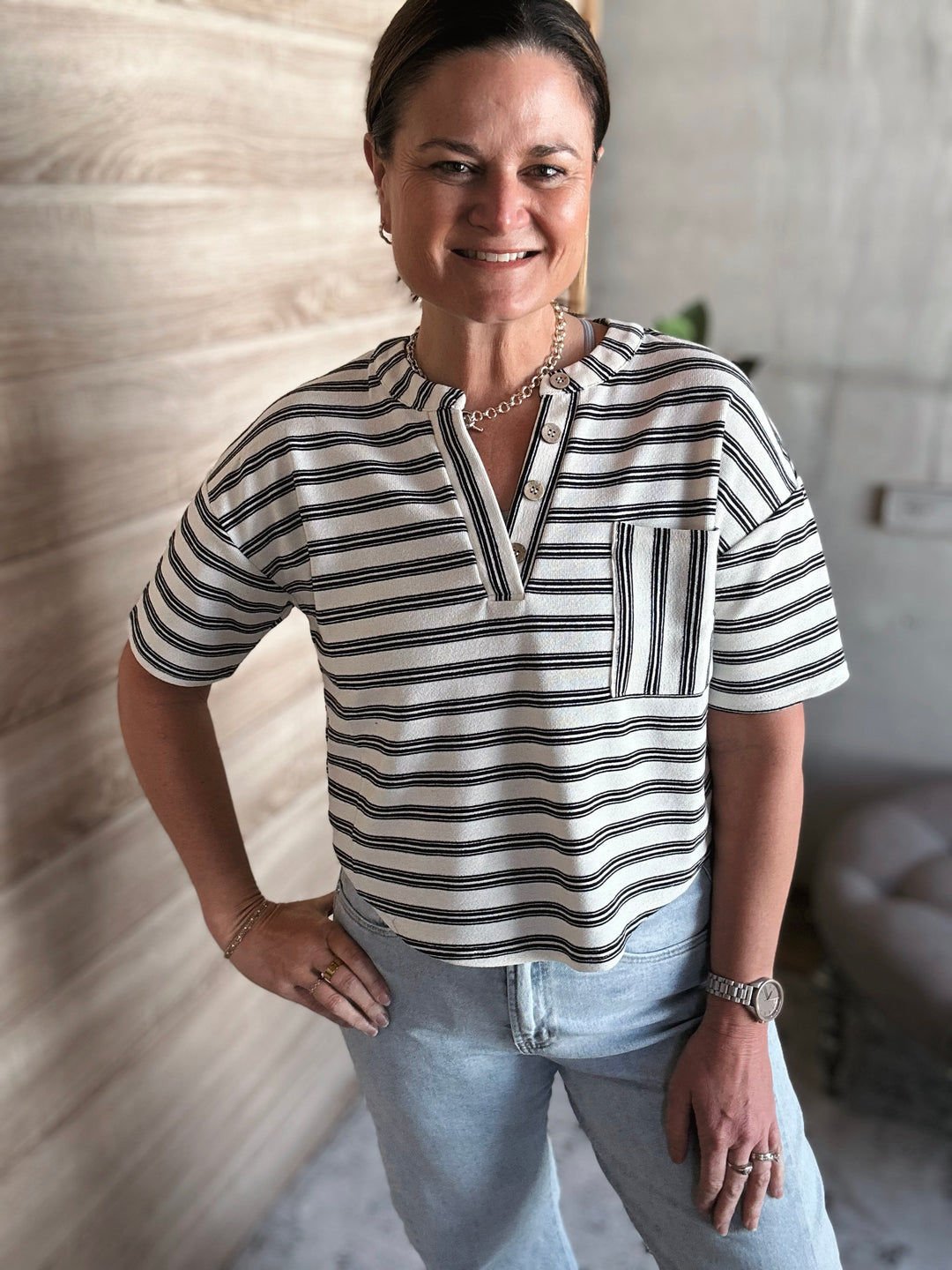 A woman wearing a short sleeved black and white striped v-neck shirt with buttons. With a pocket on the left side of the shirt. 