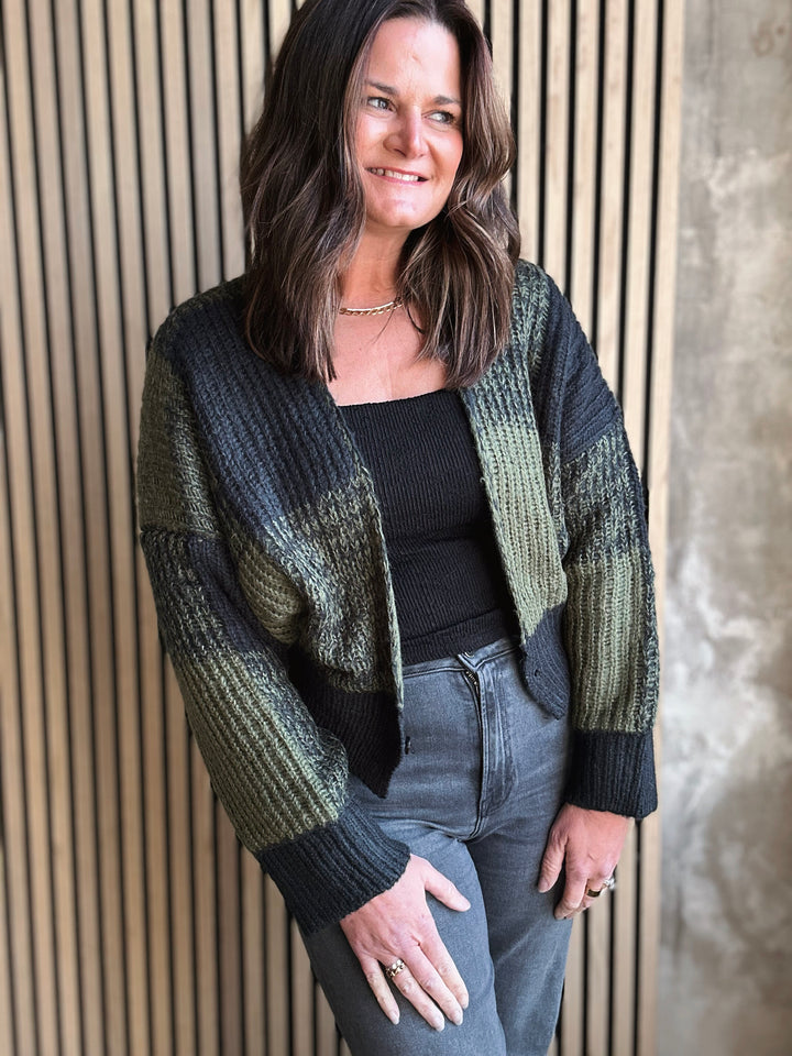 Woman wearing a green and black striped cardigan against a wooden paneled wall.