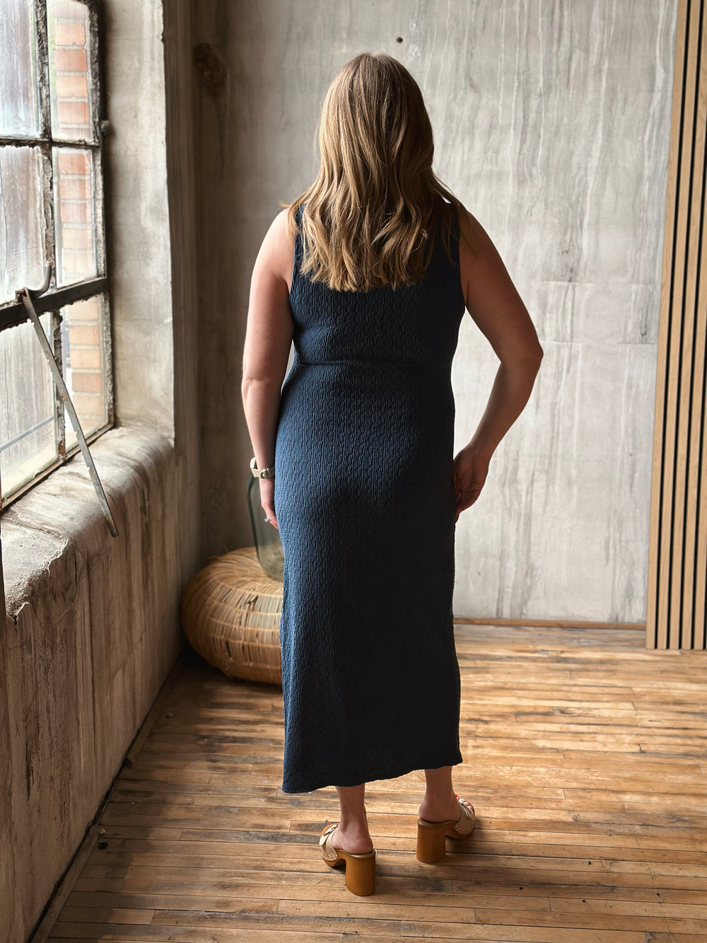Woman in a dark blue dress standing in a room with wooden floor and window.